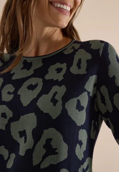 Frau in langärmeligem Shirt mit khakigrünem Leopardenmuster, lächelnd im Porträt.