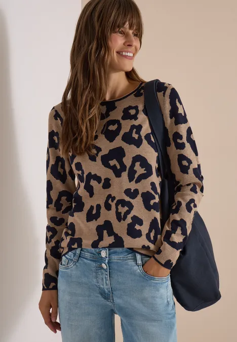 Frau mit beige-schwarzem Leo-Pullover, blauer Jeans und dunkelblauer Tasche lächelt.