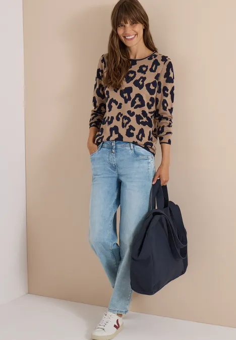 Frau lehnt an Wand in Leo-Pullover, Jeans, Sneakern und großer Tasche, sie lächelt.