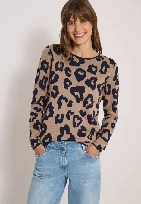 Frau lächelt in beige-schwarz getupftem Pullover und Jeans vor hellem Hintergrund.