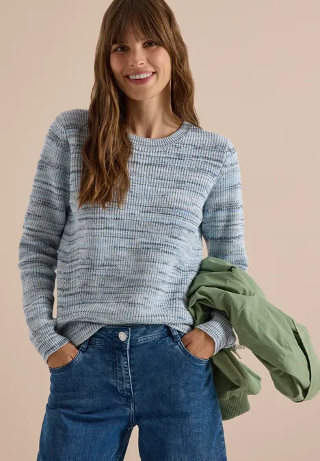 Frau mit blau-grauem Pullover, Jeans und grüner Jacke lächelt vor neutralem Hintergrund.