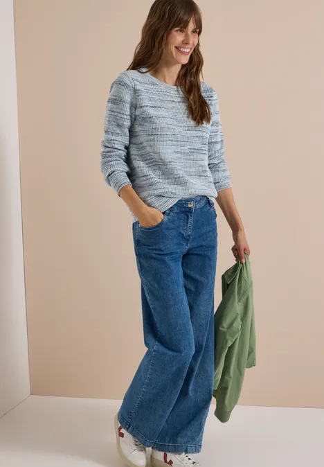 Frau in weitem Jeans, gemustertem Pullover und grüner Jacke in der Hand, lächelt vor beigefarbenem Hintergrund.