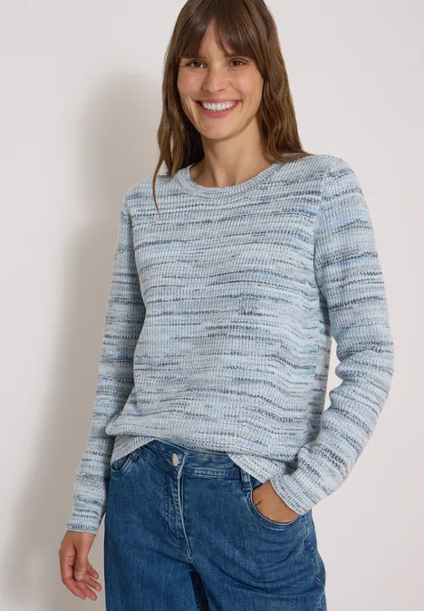 Frau mit blau-weißem Strickpullover und Jeans, lächelnd vor hellem Hintergrund.
