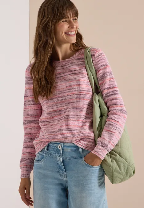Frau trägt rosa gestreiften Pullover, hellgrüne gesteppte Tasche und Jeans, lächelnd vor hellem Hintergrund.