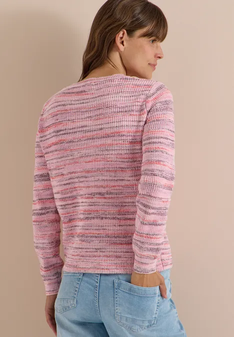 Eine Frau steht mit dem Rücken zur Kamera, trägt einen rosa gestreiften Pullover und Jeans, Hände in den Taschen.