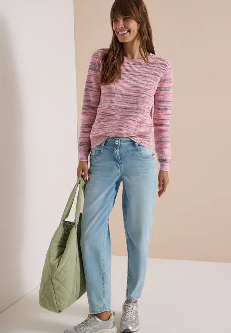 Frau in pinkem Pullover, hellblauen Jeans und Sneakern trägt eine große grüne Stepp-Tasche und lächelt.