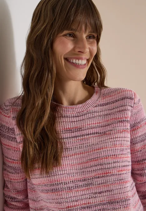 Eine lächelnde Frau mit langen, gewellten braunen Haaren lehnt an einer Wand und trägt einen rosa gestreiften Pullover.