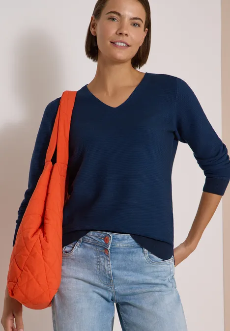 Pull bicolore à col en V urban dark blue Femme en pull bleu marine à col V, jean clair et sac orange sur l’épaule, fond clair.