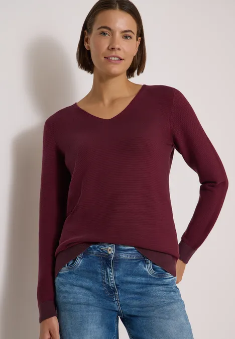 Pull bicolore à col en V berry red Une femme porte un pull bordeaux col V et un jean bleu devant un fond neutre.