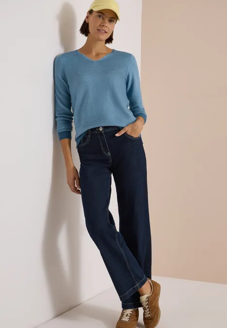Pullover im Two-Tone-Look mit V-Neck mountain air blue Frau in gelber Cap, blauem V-Pullover und dunklen Jeans lehnt an der Wand.