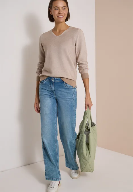 Pull bicolore à col en V creamy mocha melange Femme souriante en pull beige à col V, jean large, baskets blanches et sac vert sur fond clair.