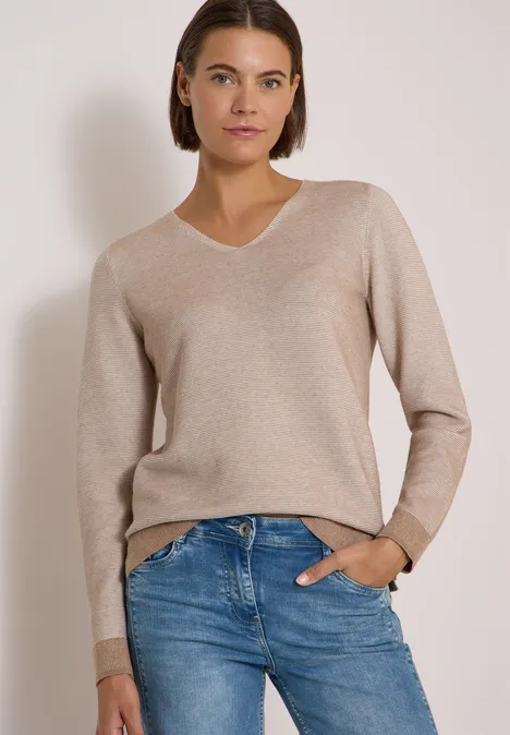 Pull bicolore à col en V creamy mocha melange Une femme aux cheveux courts bruns porte un pull beige à col V et un jean bleu clair devant un fond uni.