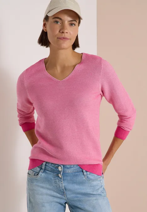 Frau mit pinkem V-Pullover, Jeans und Cap vor dezentem Hintergrund.