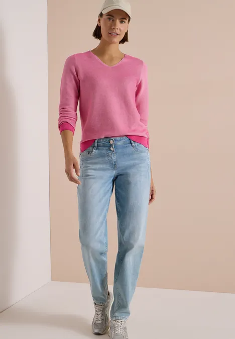 Frau in pinkem Pullover, Jeans und Cap steht vor hellem Hintergrund.