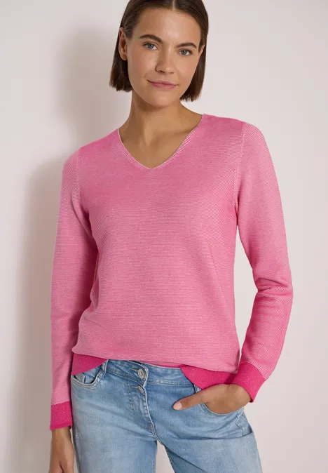 Frau trägt rosa V-Ausschnitt-Pullover und blaue Jeans, eine Hand in Tasche vor hellem Hintergrund.