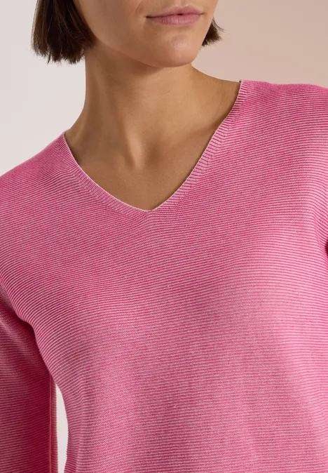 Frau trägt pinken V-Pullover, nur Oberkörper sichtbar, dezenter Hintergrund.