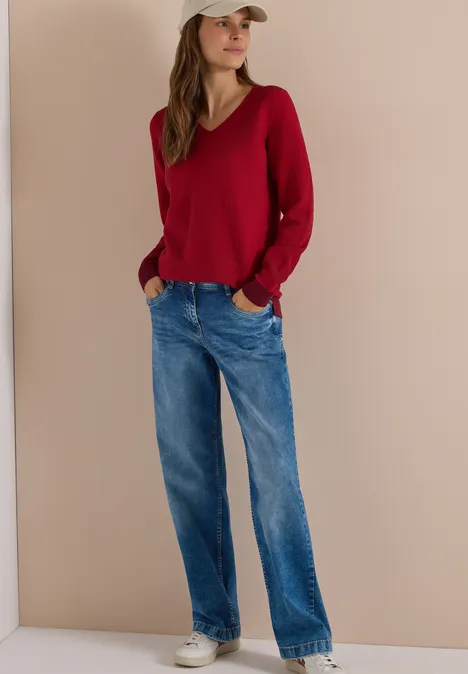 Pullover im Two-Tone-Look mit V-Neck flame red Eine Frau steht in Jeans, rotem V-Pullover, weißer Mütze und Sneakers vor beigem Hintergrund.