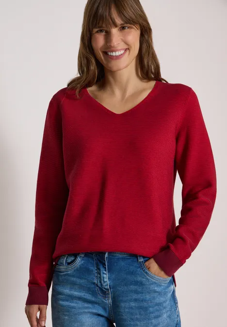 Pullover im Two-Tone-Look mit V-Neck flame red Frau mit rotem V-Ausschnitt-Pullover und Jeans, lächelnd vor hellem Hintergrund.