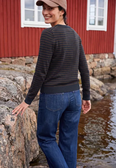 Trui met ronde hals en strepen urban dark blue melange Vrouw in jeans, gestreepte longsleeve en pet, glimlachend op rotsen voor een rood houten huis aan het water.