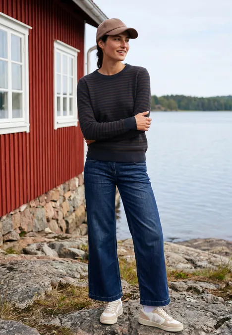 Trui met ronde hals en strepen urban dark blue melange Een vrouw met gestreepte trui, wijde jeans en pet staat op een rotsachtige oever bij een rood huis aan het meer.
