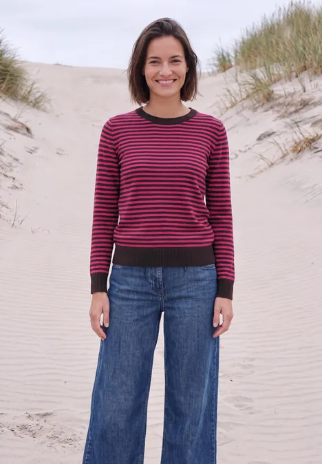 Trui met ronde hals en strepen pink flash melange Een vrouw in spijkerbroek en gestreepte trui staat op een duin bij de zee.