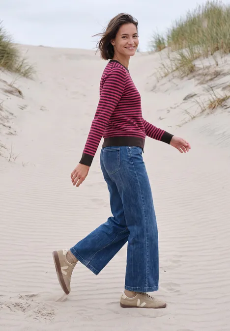 Trui met ronde hals en strepen pink flash melange Een vrouw in een rood gestreepte trui en wijde spijkerbroek loopt lachend over zandduinen op het strand.