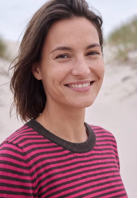 Trui met ronde hals en strepen pink flash melange Een vrouw met bruin haar in een gestreepte trui lacht blij op het strand.