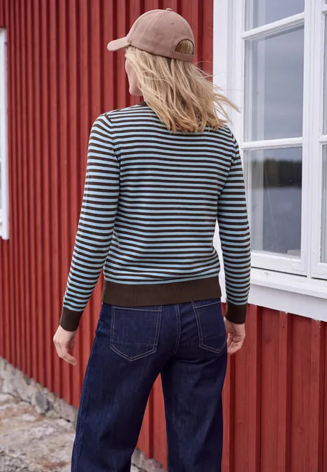 Trui met ronde hals en strepen cosmopolitan blue melange Persoon van achteren met gestreepte trui, spijkerbroek en pet voor een rood houten huis met wit raam.