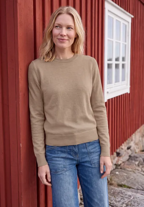 Effen trui met ronde hals creamy mocha melange Vrouw in beige trui en spijkerbroek voor een rood huis met raam.