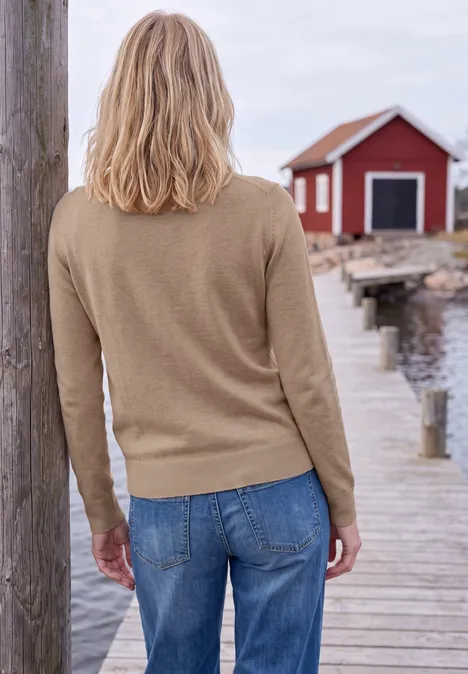Effen trui met ronde hals creamy mocha melange Persoon met blond haar in een beige trui en spijkerbroek op een houten steiger voor een rood huis aan het water.