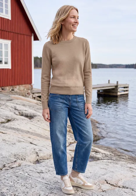 Effen trui met ronde hals creamy mocha melange Vrouw in trui, jeans en sneakers staat op een rotsachtige oever voor een rood huis aan het water