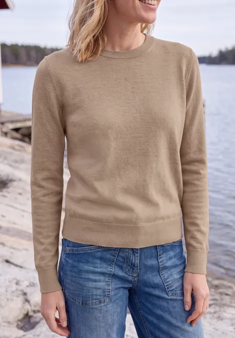 Effen trui met ronde hals creamy mocha melange Vrouw in beige trui en spijkerbroek staat bij de oever van een meer.