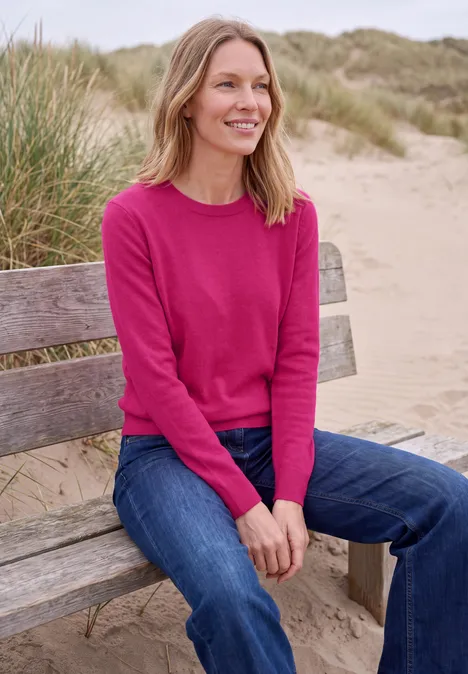 Effen trui met ronde hals pink flash melange Een vrouw zit op een bankje aan het strand, draagt een roze trui en lacht.