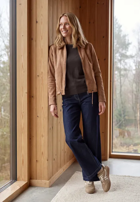 Effen trui met ronde hals truffle brown melange Een vrouw staat ontspannen bij een houten wand en raam, met een bruine jas, donker trui, blauwe jeans en beige sneakers.