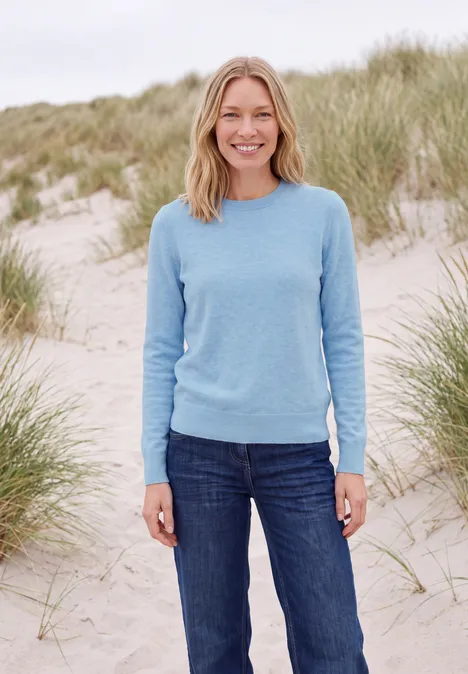 Effen trui met ronde hals cosmopolitan blue melange Een glimlachende vrouw in een blauwe trui en spijkerbroek staat op een strand tussen duinen.
