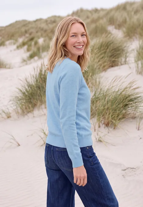 Effen trui met ronde hals cosmopolitan blue melange Lachende vrouw in blauwe trui en spijkerbroek staat op een zandstrand voor duinen.