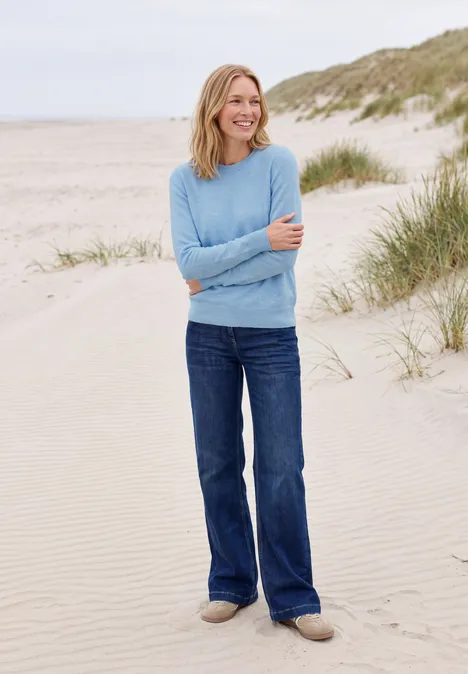 Effen trui met ronde hals cosmopolitan blue melange Een vrouw met een blauwe trui en jeans staat lachend op het strand voor duinen.