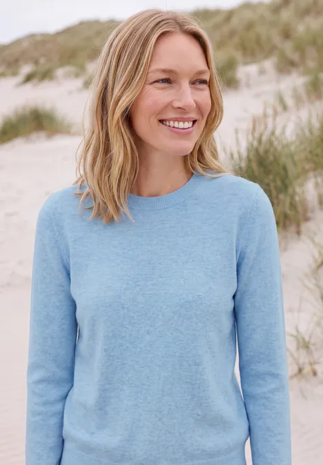 Effen trui met ronde hals cosmopolitan blue melange Vrolijke vrouw in lichtblauwe trui staand op een zandduin aan het strand.