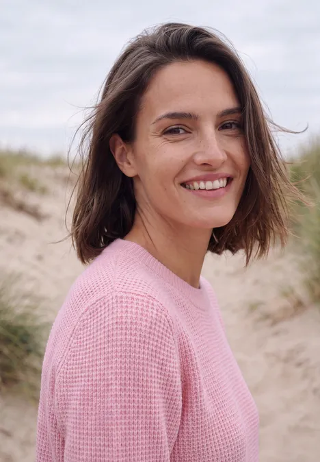 Een lachende vrouw in een roze trui op een strand met duinen op de achtergrond.