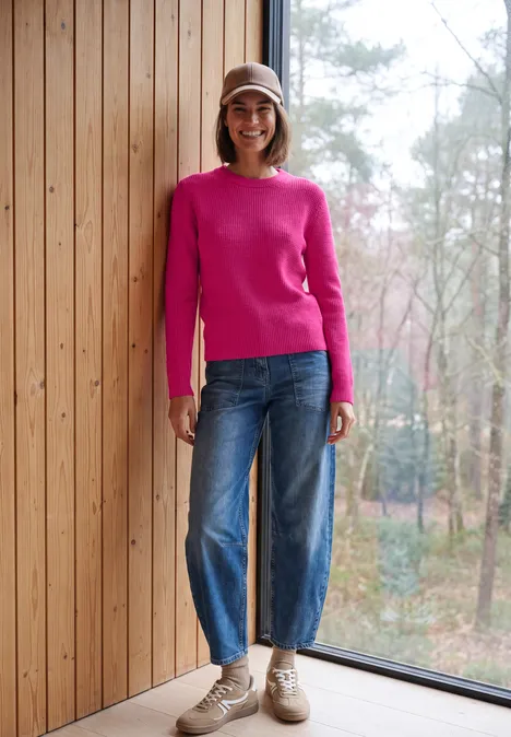 Een glimlachende vrouw in roze trui, wijde jeans en beige hoed leunt tegen een houten wand met uitzicht op natuur.