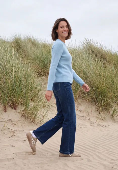 Vrouw in lichtblauwe trui en jeans loopt glimlachend over een zandstrand met helmgras op de duinen op de achtergrond.