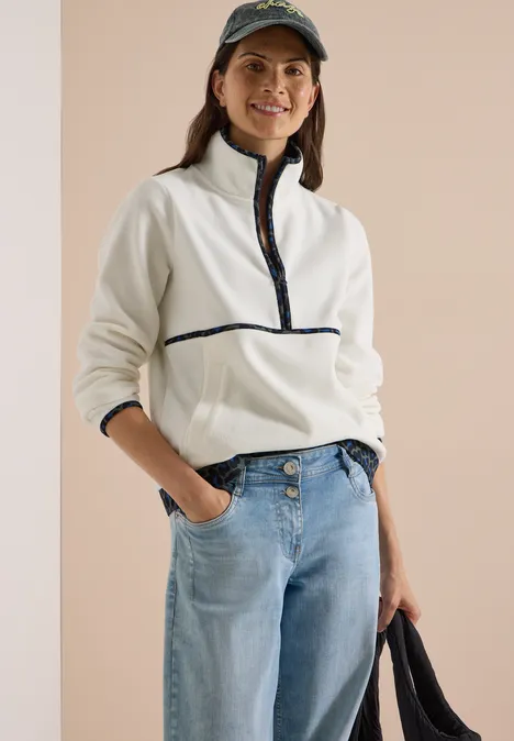 Junge Frau lächelt, trägt weiße Fleecejacke mit Reißverschluss, Jeans und Cap, hält eine Tasche.