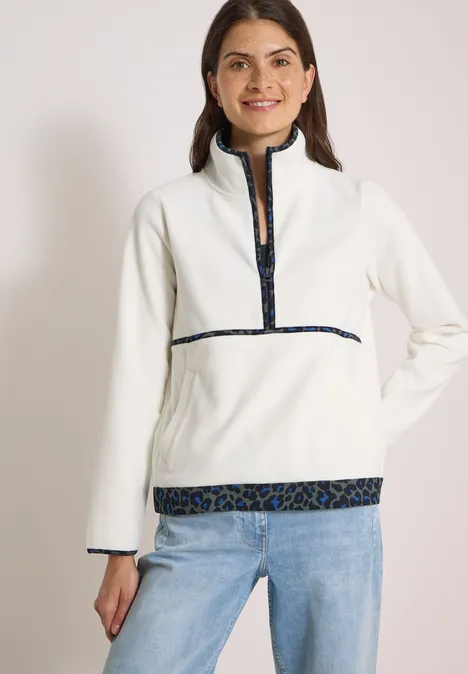 Eine Frau in weißem Fleece-Pullover mit halbem Reißverschluss und gemusterten Abschlüssen.