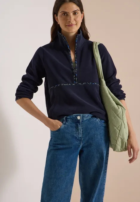 Fleecetrui met leopardetails urban dark blue Vrouw in marineblauw fleece shirt met rits, blauwe spijkerbroek en groene schoudertas, recht vooruit kijkend.