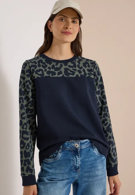 Vrouw in marineblauwe trui met luipaardprint op de mouwen, spijkerbroek en pet, voor neutrale achtergrond.