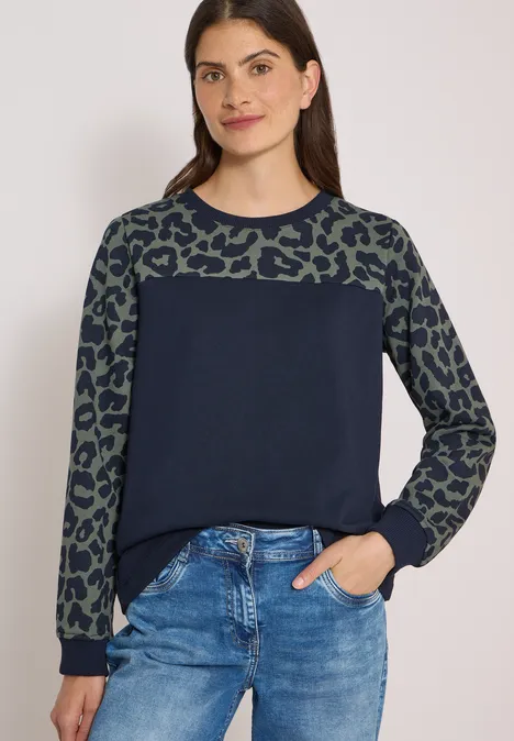 Vrouw in jeans en marineblauwe trui met groene luipaardprintmouwen, hand in de zak.