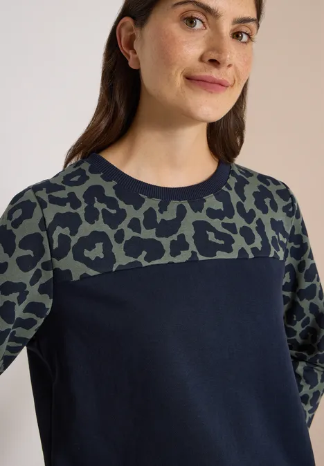 Vrouw in een donkerblauw sweatshirt met groen-zwart luipaardprint op borst en mouwen.