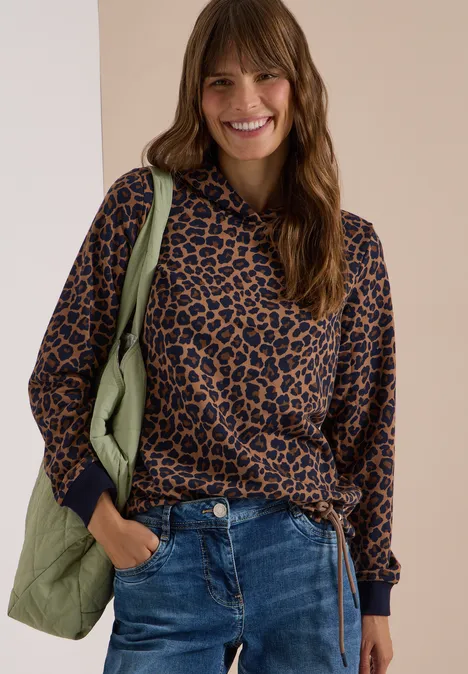 Kauzige Frau in Leoparden-Hoodie mit Jeans und grüner Tasche lächelt.