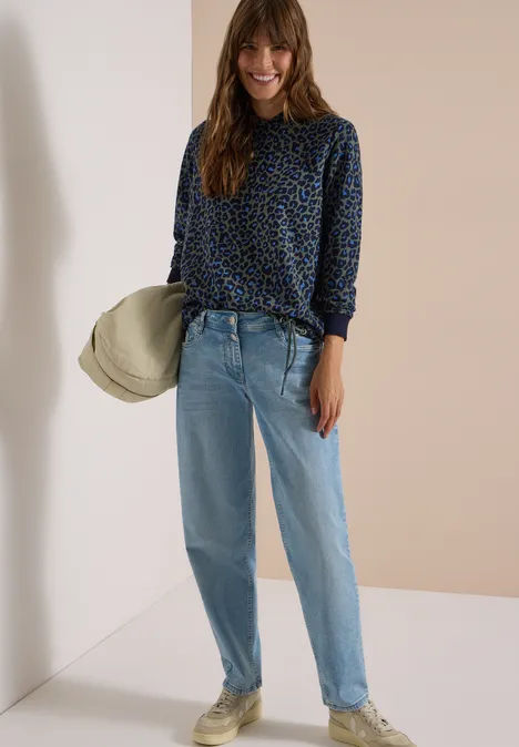 Frau mit gemusterter Bluse, hellen Jeans und Hut in neutraler Umgebung.