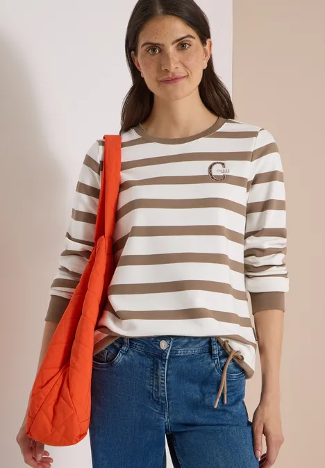Frau mit weiß-beige gestreiftem Langarmshirt, Jeans und oranger Tasche vor hellem Hintergrund.